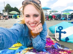 Blonde 20-something girl laying on stomach on beach, with one hand folded under her chin and the other taking a selfie. Sunglasses are on her head and a colorful beach towel is underneath her.