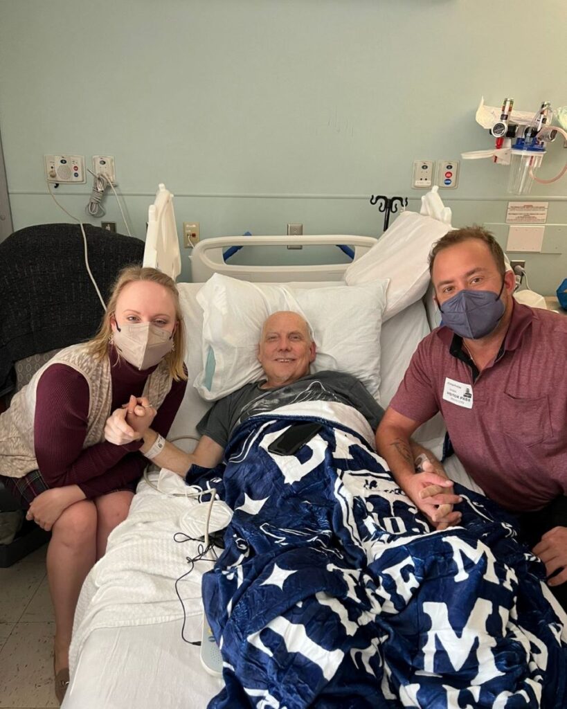 A young woman in her 30s wearing a mask sits next to her father who is laying on a hospital bed. On the other side of him is a young man in his 40s. Both children are holding one of their father's hands as he lays between them.